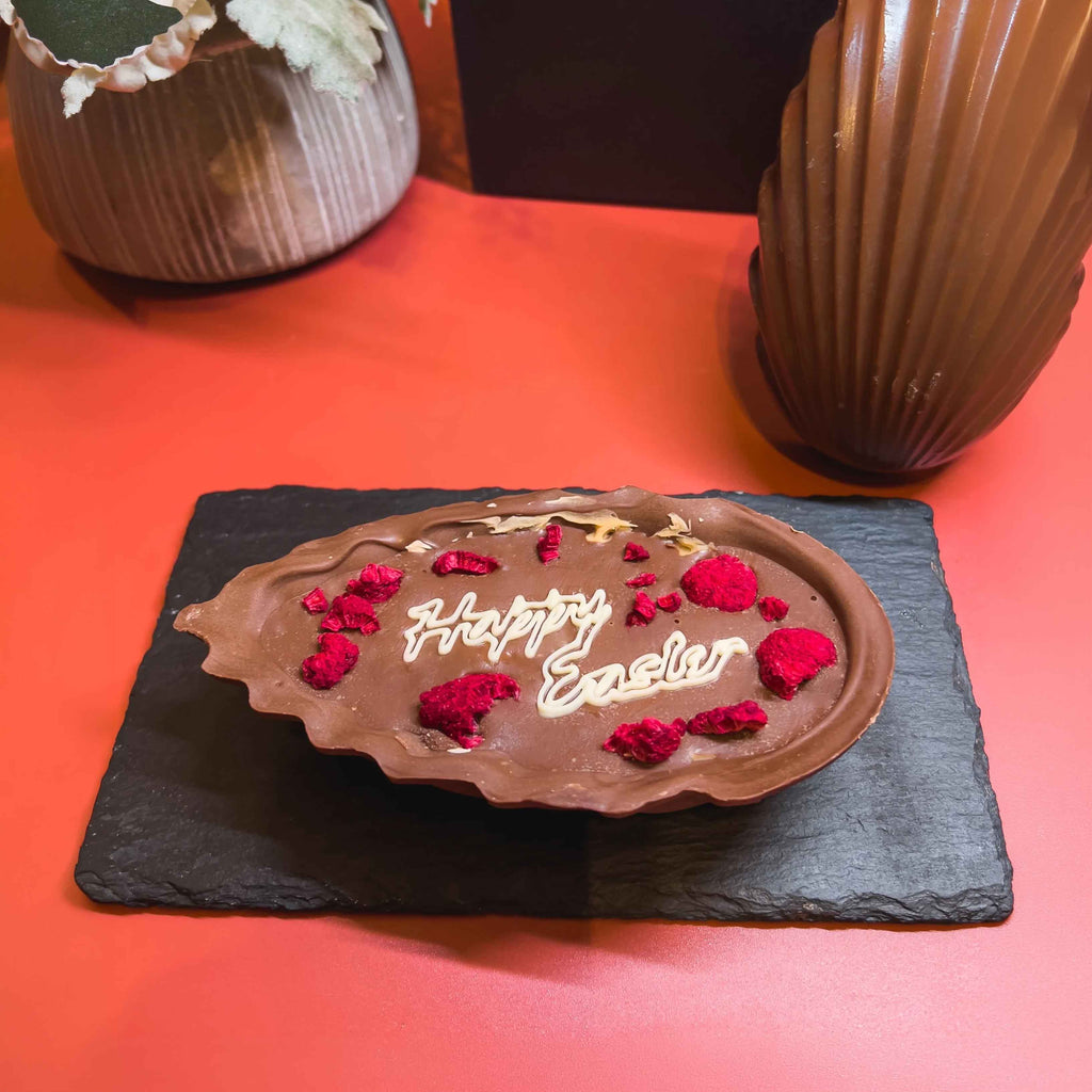 Signature Easter Egg: filled milk chocolate half topped with dried raspberries and Happy Easter in white chocolate, beside the hollow half