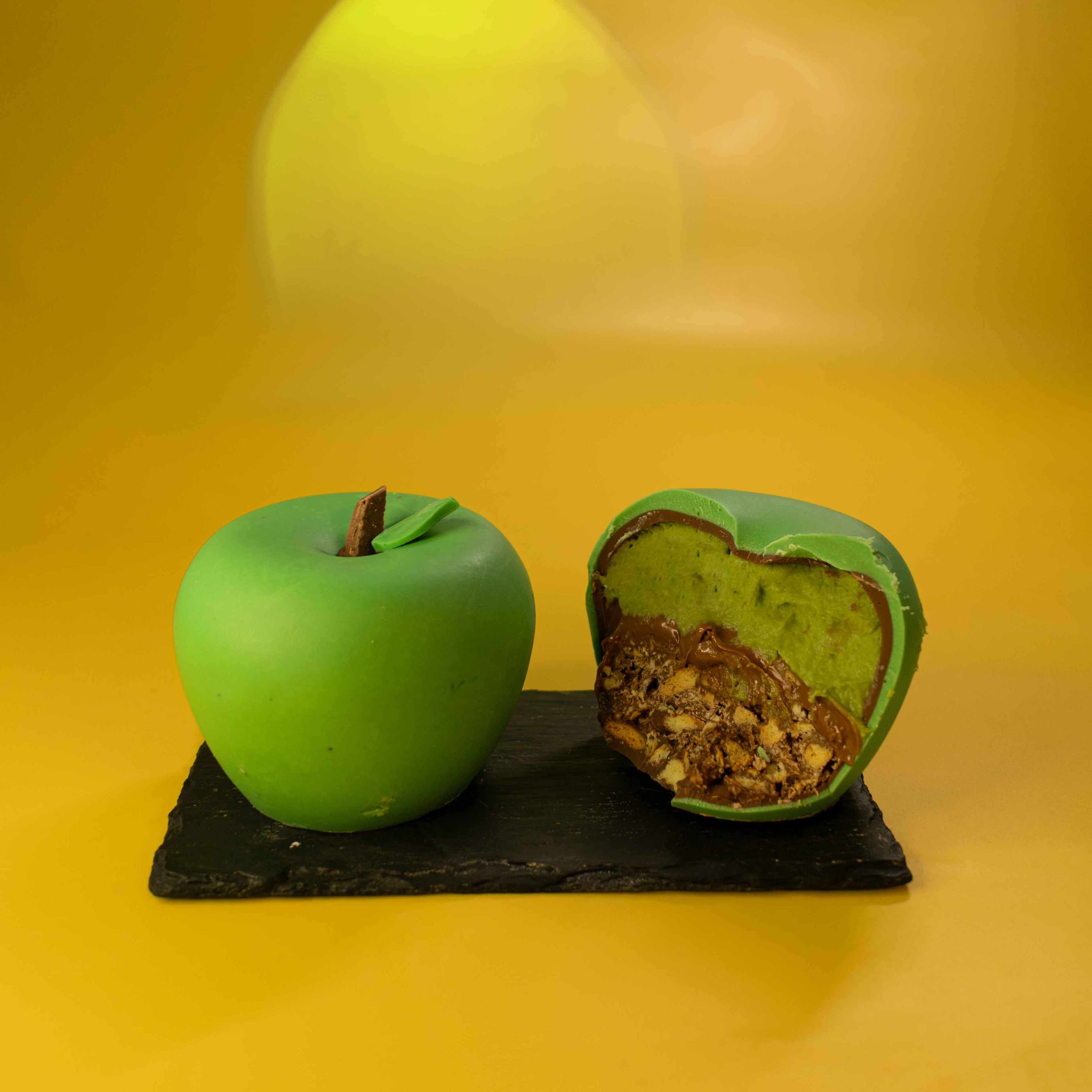 A green apple-shaped confection sits on a black slate board against a warm yellow background. One whole apple-shaped piece is shown on the left, and a cut piece on the right reveals a green outer layer and a nut-filled interior.